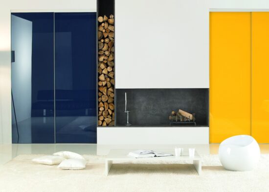 Un salon moderne au design élégant et minimaliste avec des portes bleues et jaunes audacieuses, une cheminée centrale avec un coin de rangement en bois et un mobilier contemporain élégant sur fond de murs et de cuisine blancs.