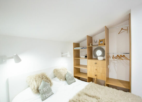 Une chambre confortable et minimaliste avec un lit soigneusement fait avec des oreillers « joyeuses fêtes », une tête de lit en bois avec étagères intégrées et une applique murale blanc chaud sous rampante.
