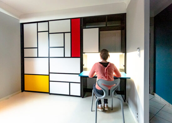 Un enfant est assis à un bureau moderne dans une pièce bien éclairée avec une étagère colorée et géométrique au mur, accompagnée de panneaux publicitaires vibrants.