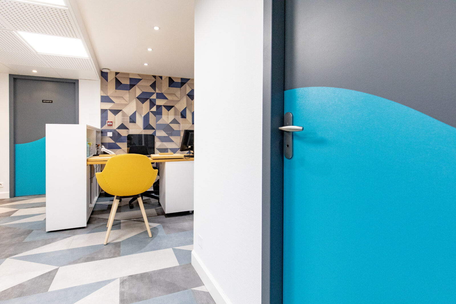 Un espace de bureau moderne et élégant avec des motifs géométriques, avec un mur et une porte aux accents bleu vif, une chaise jaune élégante et un sol à motifs offrant un environnement de travail frais et contemporain.