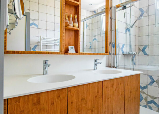 Une salle de bain lumineuse et aérée comprenant une vanité en bois avec un comptoir blanc, un grand miroir et une douche vitrée avec des carreaux bleus et blancs.