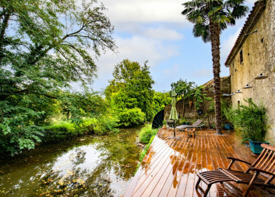 Le charme rustique rencontre la tranquillité naturelle : une terrasse Annie sereine au bord de la rivière avec une terrasse en bois et des chaises relaxantes à côté d'une vieille maison en pierre, au milieu d'un feuillage verdoyant et sous un ciel clair et nuageux.