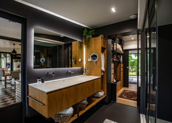 Une salle de bains moderne et élégante avec des accents en bois et des plantes suspendues, dotée d'une double vasque, d'un grand miroir et d'une vue sur une pièce adjacente à travers des vitres après une restauration complète.
