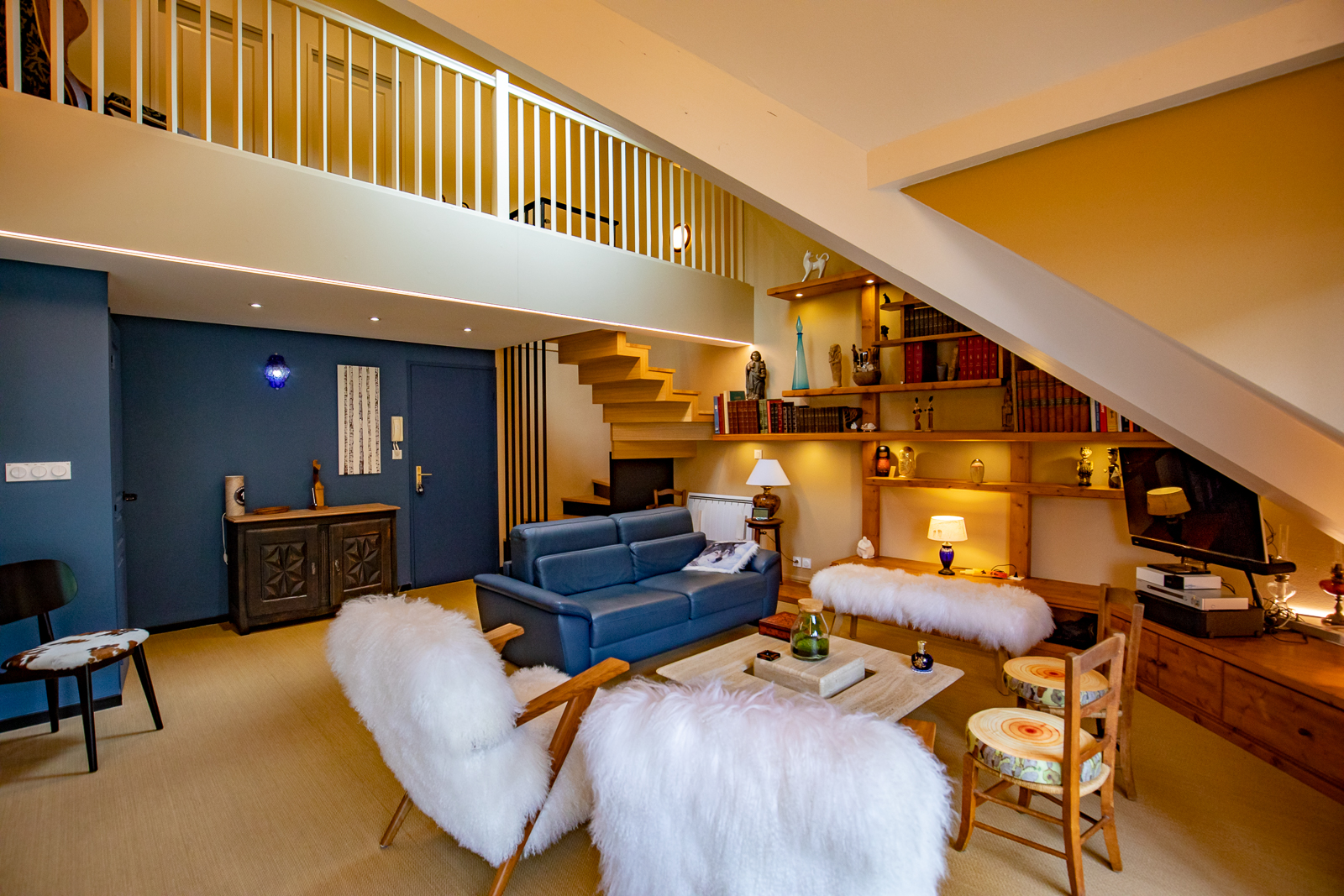Un salon cosy et moderne dans un agencement total d'appartements avec un canapé bleu moelleux, des fauteuils blancs moelleux et une ambiance lumineuse chaleureuse, complétée par une mezzanine avec