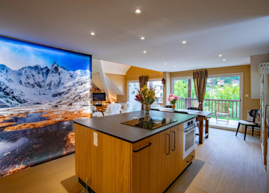 Une cuisine moderne avec un grand îlot central faisant face à une superbe photographie murale d'un paysage de montagne enneigé, complétée par la lumière naturelle entrant depuis le balcon adjacent.