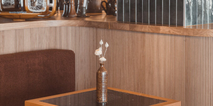 Un coin douillet dans un café élégant aux tons chauds de bois et à la décoration élégante, comprenant une petite table dressée avec un vase de fleurs pittoresque, flanquée de chaises accueillantes, prête à accueillir les invités pour un moment de détente.