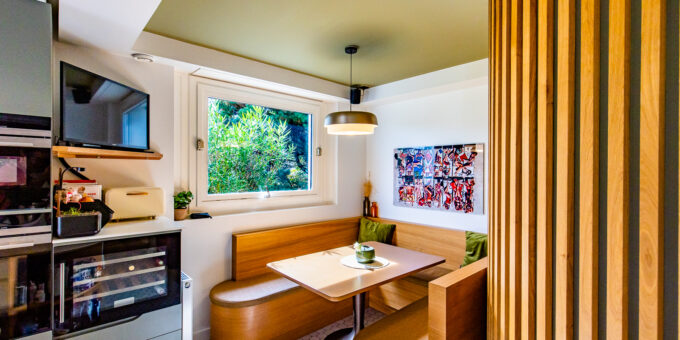 Un coin cuisine moderne et confortable avec des sièges intégrés, des accents en bois et des œuvres d'art vibrantes, baigné de lumière naturelle depuis une fenêtre donnant sur la verdure, conserve le cachet d'un 70