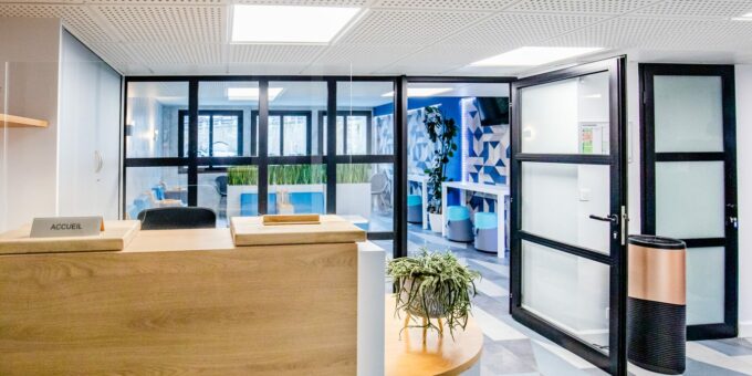 Espace de réception de bureau moderne conçu par un architecte d'intérieur à Tarbes, avec un bureau en bois épuré, une décoration végétale et un aperçu de l'espace lounge stylé à travers les portes vitrées.