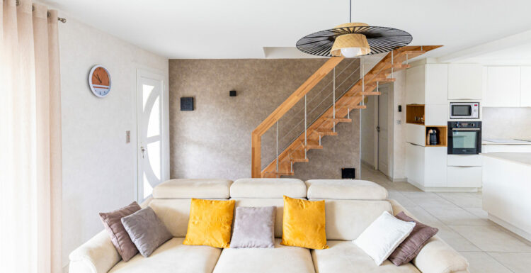 Un salon lumineux et moderne avec un canapé blanc orné d'oreillers jaunes et violets présente un ventilateur de plafond unique. La pièce comprend un escalier, des appareils de cuisine et est idéale pour tous ceux qui recherchent "sér