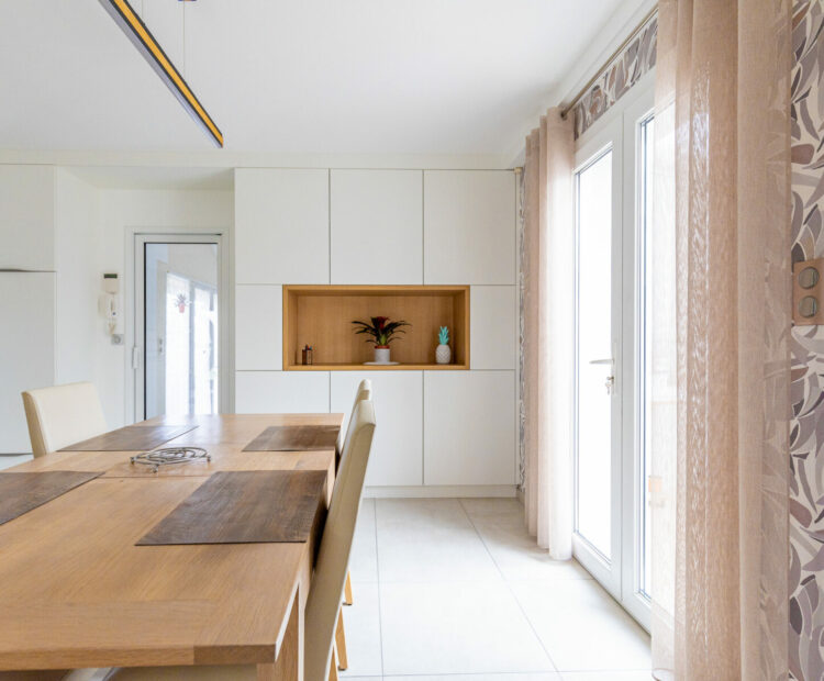 Une cuisine lumineuse et moderne avec des murs blancs, une grande table à manger en bois et des armoires encastrées conçues pour la sérénité. La lumière naturelle pénètre à travers une porte vitrée et le décor comprend un minimum de