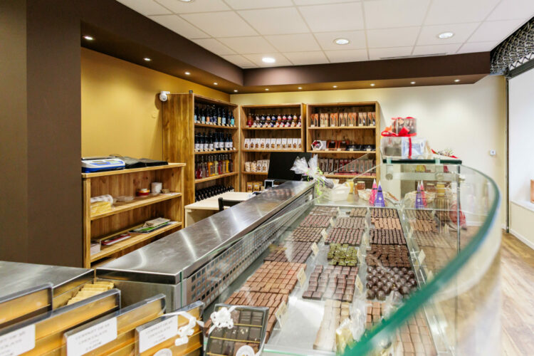 Un intérieur de chocolaterie bien éclairé, un joli agencement intérieur, avec des étagères présentant une variété de produits chocolatés à gauche et une vitrine en verre au premier plan présentant un assortiment de chocolats et de truffes. Une caisse enregistreuse est visible sur un comptoir derrière le présentoir.