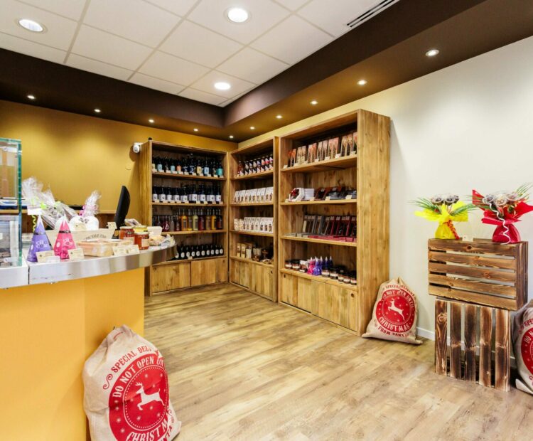 Une chocolaterie cosy avec des étagères en bois présentant divers produits chocolatés. Le comptoir de gauche est doté d'un écran d'ordinateur et est décoré d'articles cadeaux emballés. Le sol est en bois et l’éclairage est chaleureux, créant une atmosphère accueillante, grâce à notre équipe dédiée projets d’agencement intérieur.