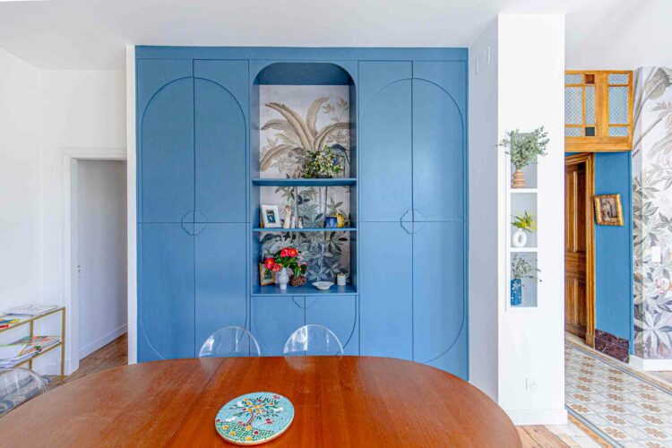 Une salle à manger avec une table en bois et des chaises transparentes. L'arrière-plan présente une grande armoire bleue avec deux sections fermées et une section centrale ouverte présentant des plantes, une photo encadrée et des objets décoratifs. Les murs présentent un décor coloré et à motifs, reflétant l'agencement intérieur minutieux de notre équipe.