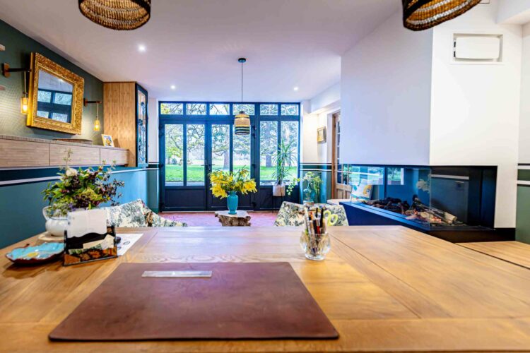 Un bureau cosy avec une grande table en bois au centre, ornée d'un sous-main en cuir et d'articles de papeterie. L'espace comprend des fauteuils fleuris, une cheminée moderne, un éclairage élégant et des baies vitrées qui révèlent une vue extérieure verdoyante