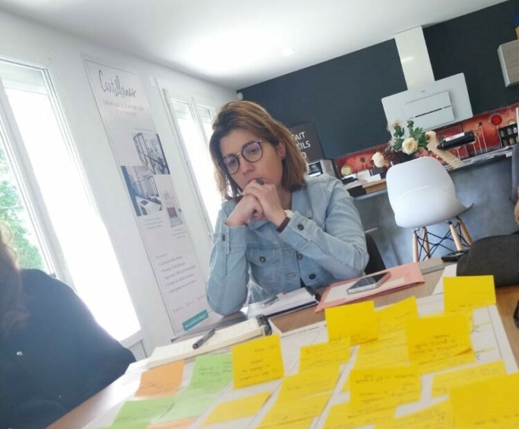 Une personne portant des lunettes et une veste en jean est assise à une table recouverte de notes autocollantes et de papiers colorés. Ils semblent plongés dans leurs réflexions, peut-être en train de réfléchir ou de travailler sur un projet d'architecture d'intérieur, dans une pièce lumineuse avec de grandes fenêtres et une cuisine moderne en arrière-plan.