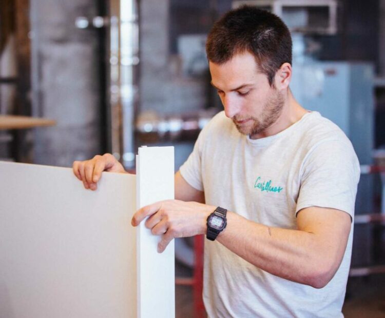 Un homme portant un T-shirt blanc et une montre-bracelet travaille sur un projet de menuiserie. Il assemble un meuble en maintenant deux panneaux blancs ensemble sur un établi. Divers outils et matériaux sont disséminés autour de l’établi, rappelant un entretien de Léa avec une stagiaire en architecture d’intérieur.