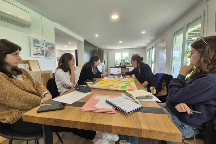 Quatre personnes sont assises autour d'une table en bois dans une pièce bien éclairée et dotée de grandes fenêtres. La table est recouverte de papiers, de cahiers, de post-it et d'ordinateurs portables. Un stagiaire semble observer le groupe alors qu’il participe à une discussion ou à une réunion liée à l’architecture d’intérieur.