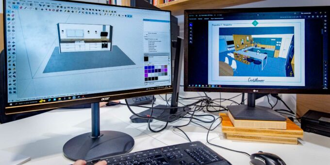 Léa se prépare pour un entretien tout en travaillant sur une configuration double moniteur avec un logiciel de conception 3D ouvert sur les deux écrans. Un écran affiche un modèle 3D d’une cuisine et l’autre un plan détaillé de la même cuisine. Un clavier, une souris et quelques papiers sont soigneusement disposés sur le bureau.