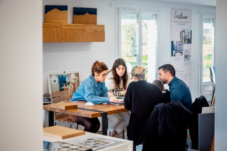 Quatre personnes, dont Léa, sont assises autour d'une table dans un bureau lumineux. Ils consultent des documents et discutent intensément, peut-être pour un entretien sur l’architecture d’intérieur. Le groupe semble concentré et engagé. Le bureau dispose de grandes fenêtres, d’œuvres d’art sur des étagères et d’une bannière en arrière-plan.