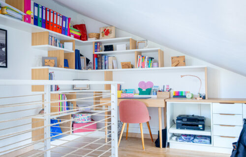 Bureau à domicile mansardé lumineux et confortable comprenant un bureau en bois, une chaise rose et des étagères de rangement blanches remplies de classeurs colorés, de livres et d'objets de décoration. Un plafond incliné ajoute du caractère et un étendoir à linge se trouve au premier plan : le refuge créatif parfait de Barbara.