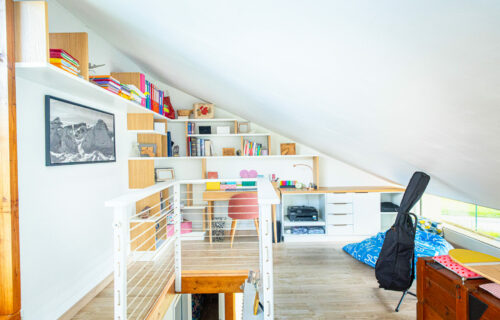 Une chambre mansardée lumineuse avec un plafond incliné dispose d'un parquet, d'un bureau avec une chaise rose et d'étagères remplies de livres et d'objets divers. Une photo encadrée de Barbara est suspendue au-dessus du bureau en forme de bureau. Une guitare sur pied et une couverture à motifs bleus sont visibles au sol.