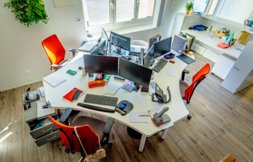 Un espace de bureau bien éclairé avec quatre bureaux incurvés disposés en cercle, chacun mettant en valeur son expertise avec un écran d'ordinateur et des fournitures de bureau. Les chaises rouges ajoutent une touche d'Adour au parquet. Une plante prospère dans un coin près d’une grande fenêtre et des étagères sont visibles à l’arrière-plan.