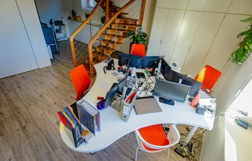 Un bureau moderne d'Adour Expertise comprend un bureau blanc de forme unique avec plusieurs écrans d'ordinateur, claviers et fournitures de bureau. Le bureau est entouré de chaises rouges sur un sol à motifs de bois. Un escalier en bois et des plantes en pot ajoutent une touche de décoration naturelle à cet espace lumineux.