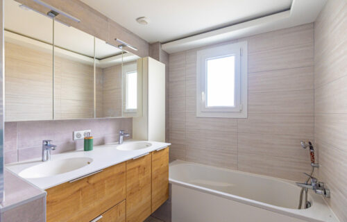 Une salle de bain moderne dotée d'une vanité en bois avec double vasque et de grands miroirs au-dessus. Une fenêtre illumine l'espace de lumière naturelle. La baignoire, équipée d'une pomme de douche, se trouve à droite du meuble-lavabo. Les murs sont carrelés dans des tons gris clair, ajoutant de l'élégance au design Gille.