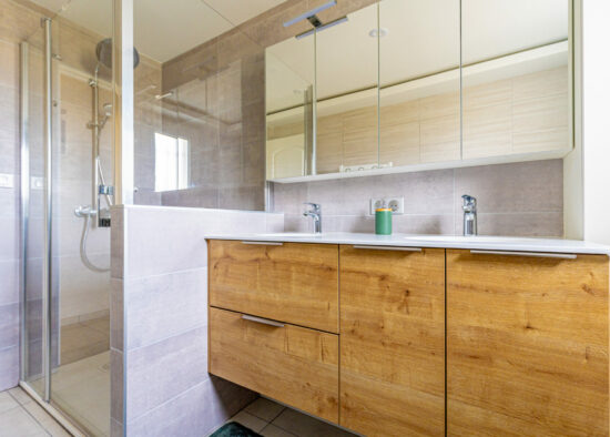 Une salle de bain moderne avec du carrelage beige clair et une douche vitrée. Il y a une vanité en bois avec deux lavabos et robinets chromés, surmontée d'un grand miroir avec armoires. L'espace est lumineux et contemporain, offrant à la fois un style et une fonctionnalité semblables aux créations de Gille.