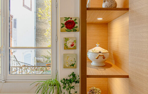 Un coin cuisine lumineux avec un meuble en bois présentant de la porcelaine décorative. Le meuble comporte des étagères éclairées présentant diverses théières et bocaux à motifs floraux. Un comptoir avec des herbes en pot se trouve à côté d'une fenêtre, offrant une vue sur les bâtiments voisins, mettant en valeur l'essence de la cuisine française.