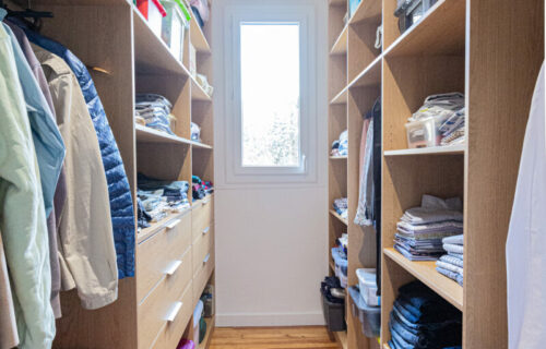 Un dressing avec des étagères en bois des deux côtés, soigneusement organisé avec des vêtements pliés, des boîtes et divers objets. Les manteaux et les vestes sont suspendus sur des cintres à gauche. Une valise rouge est visible en bas à gauche. Une fenêtre au fond apporte de la lumière naturelle à cet élégant dressing pour Constance.