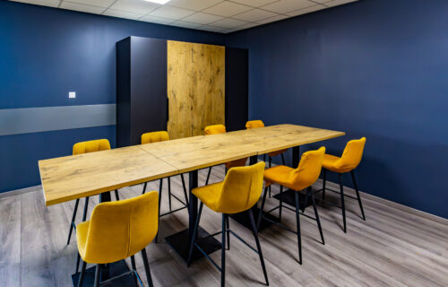 Une salle de conférence moderne aux murs bleu foncé et au parquet conçue par Dupuy Agencement. Il comporte une grande table rectangulaire en bois entourée de chaises rembourrées jaune vif. Il y a un panneau décoratif en bois sur le mur et un interrupteur à LED conçu avec des détails méticuleux.