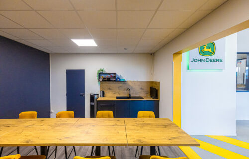 Une kitchenette de bureau moderne et un espace de réunion. L'espace comprend une table en bois avec des chaises orange, un petit comptoir de cuisine avec four micro-ondes et bouilloire, ainsi qu'une porte. Les murs bleus et blancs présentent un agencement de Dupuy, présentant un grand logo John Deere à côté d'une porte menant à une autre pièce.