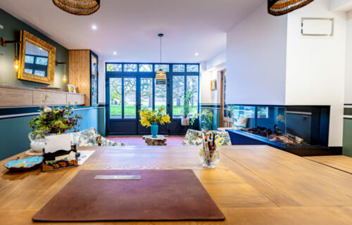 Un salon cosy et bien éclairé avec une grande table en bois du Bureau Trémont au premier plan, une cheminée à droite et des portes françaises donnant sur un espace jardin. La pièce dispose de sièges confortables, de fleurs jaune vif et de divers éléments décoratifs comme des suspensions et un miroir encadré.