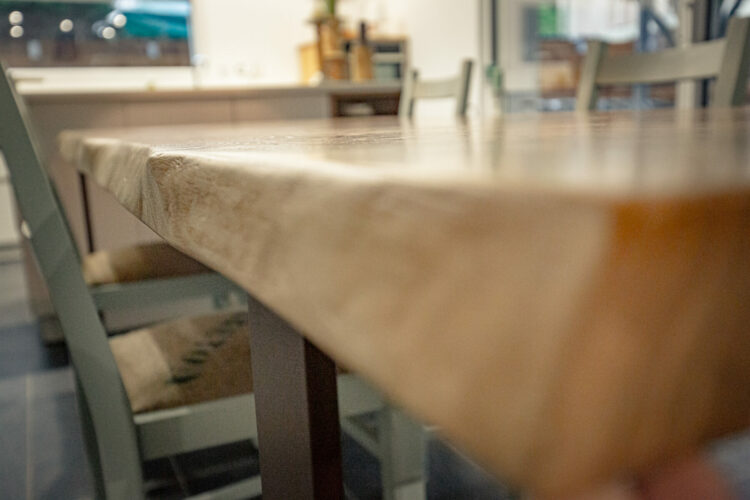 Gros plan sur une table à manger en bois aux finitions lisses dans une cuisine lumineuse, rappelant un élégant projet d’agencement. Un éclairage doux met en valeur la texture du bois. En arrière-plan, des images floues de chaises et d’éléments de cuisine créent une scène accueillante digne de toute maison d’Auch.