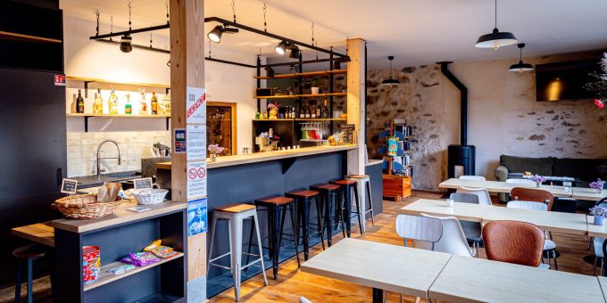 Un intérieur de café chaleureux avec un sol en bois et un mur en pierre. Cet espace multifonctionnel dispose d'un bar noir avec de hauts tabourets, d'une sélection soignée de boissons, d'étagères avec des produits Seich et d'un panier de collations. Plusieurs tables avec diverses chaises peuplent la salle à manger sous d'élégantes suspensions.