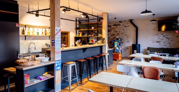 Un intérieur de café chaleureux avec un sol en bois et un mur en pierre. Cet espace multifonctionnel dispose d'un bar noir avec de hauts tabourets, d'une sélection soignée de boissons, d'étagères avec des produits Seich et d'un panier de collations. Plusieurs tables avec diverses chaises peuplent la salle à manger sous d'élégantes suspensions.